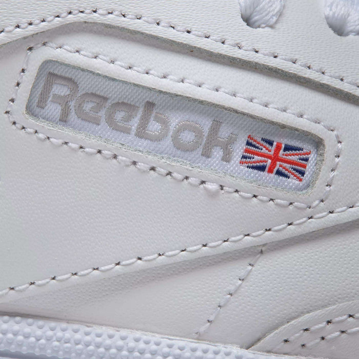 Close-up of Reebok Women's Club C 85 Shoes in white, featuring a stitched Reebok logo and Union Jack flag on a leather panel with visible stitching—showcasing iconic style and detailed craftsmanship.