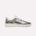 A side view of the Reebok Women’s Club C 85 Shoes in Silver Metallic/Chalk/New Grey 1 shows off their textured metallic leather upper and crisp white sole against a plain light background. Inspired by classic tennis style, this design combines comfort with eye-catching shine.