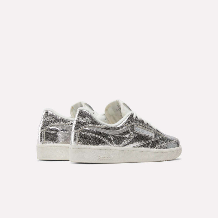 A pair of Reebok Women’s Club C 85 Shoes in metallic silver leather, featuring white soles, are positioned side by side and angled slightly away on a plain light background.