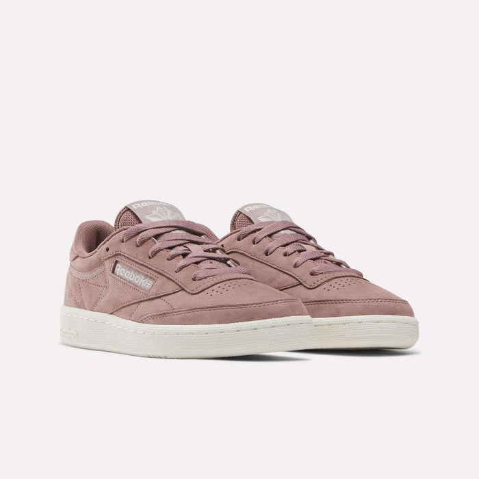 A pair of Reebok Women’s Club C 85 Shoes featuring mauve suede uppers, white soles, and branded tongues is displayed on a white background.