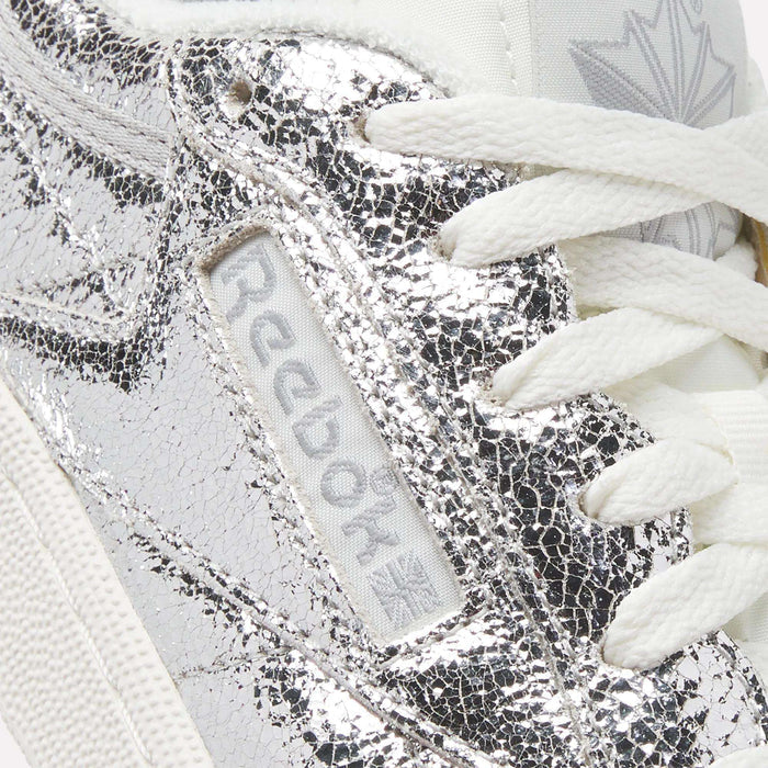 Close-up of a silver cracked-texture metallic leather Women’s Club C 85 Shoe by Reebok, featuring white laces, the Reebok logo tag on the side, and part of the maple leaf logo visible on the tongue.