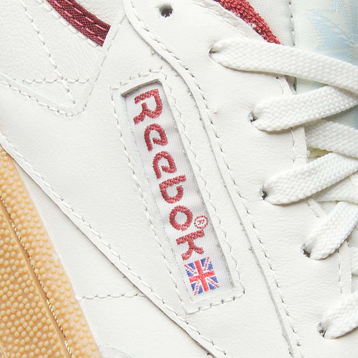 Close-up of Reebok Women's Club C Golf Shoes, showing white water-resistant leather, a stitched red logo with Union Jack flag, white laces, and a beige textured sole for enhanced durability.
