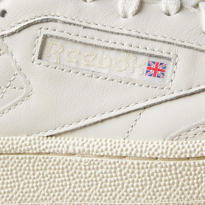Close-up of Women's Club C Golf Shoes by Reebok in white water-resistant leather, featuring the Reebok logo, a small Union Jack flag on the side, fine stitching, and a spikeless, textured golf shoe-inspired sole.