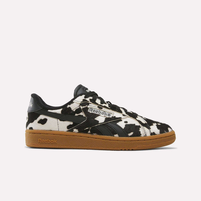 A pair of Reebok Women’s Club C Grounds UK 1.0 Shoes in Black/Brown/Gum is displayed from the side against a plain light background, highlighting their eye-catching black and white cow print pattern with sleek black accents and a brown gum sole. These animal print sneakers offer a stylish update to the iconic Reebok silhouette.