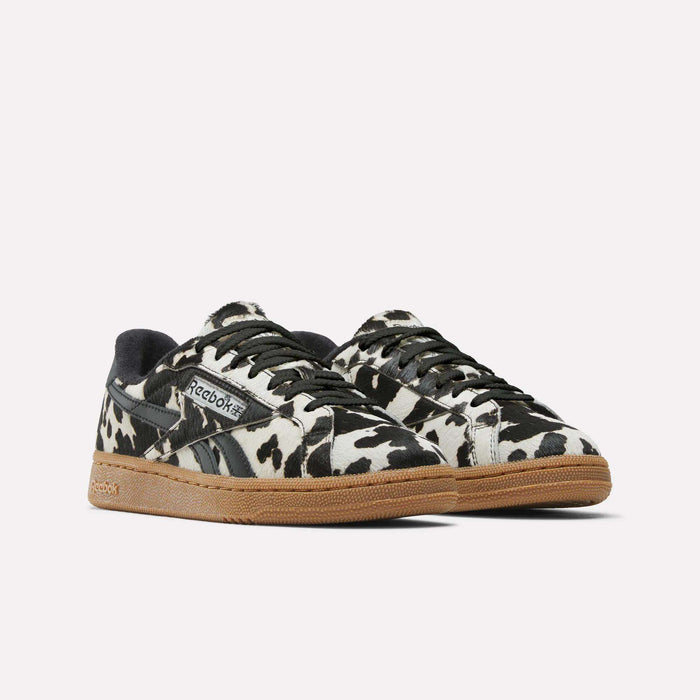 The Reebok Women's Club C Grounds UK Shoes feature a striking black and white cow print, black accents, and light brown gum soles, shown against a plain light background.