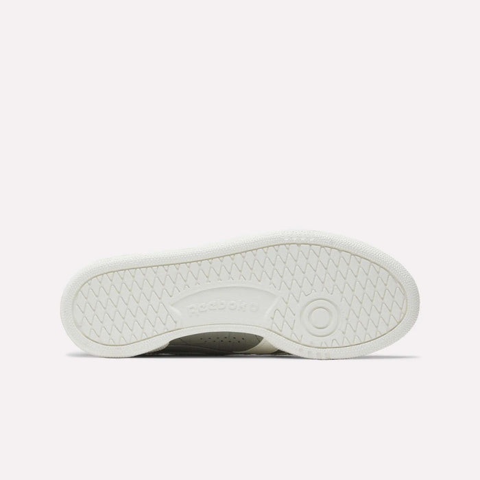 The image displays the white rubber outsole of Reebok Women's Club C Grounds UK Shoes, with a circular tread near the heel, herringbone traction patterns, and an embossed Reebok logo at the center.