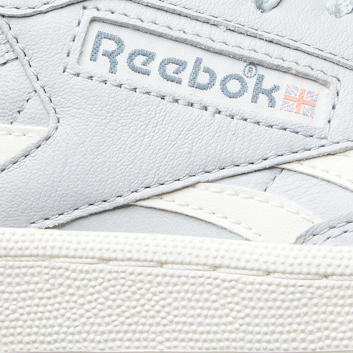 Close-up of a white Reebok Women’s Club C Revenge Shoe, highlighting its vintage court style. The iconic Reebok logo and a small British flag appear on a fabric label stitched onto the leather upper, while the textured midsole accentuates its classic design—making these casual shoes from Reebok ideal for everyday wear.