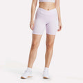 A woman models Reebok Women's Core High-Rise Cycle Shorts in light purple with a crossover waistband, paired with a matching crop top, white ankle socks, and sneakers against a plain white background.