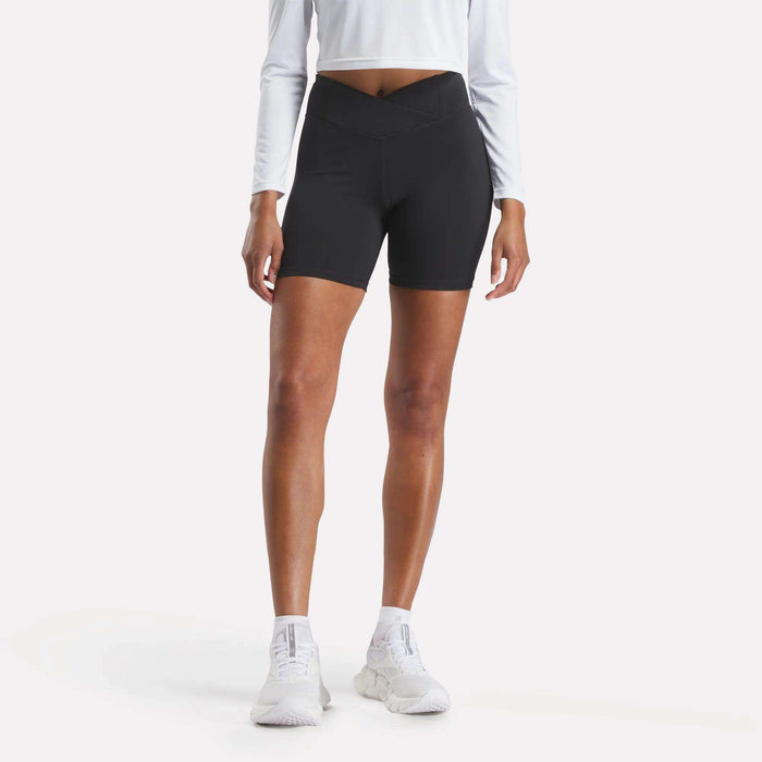 A person stands against a plain light background wearing a long-sleeve white crop top, white athletic shoes, and Reebok Women's Core High-Rise Cycle Shorts in black. The person's head is not visible.