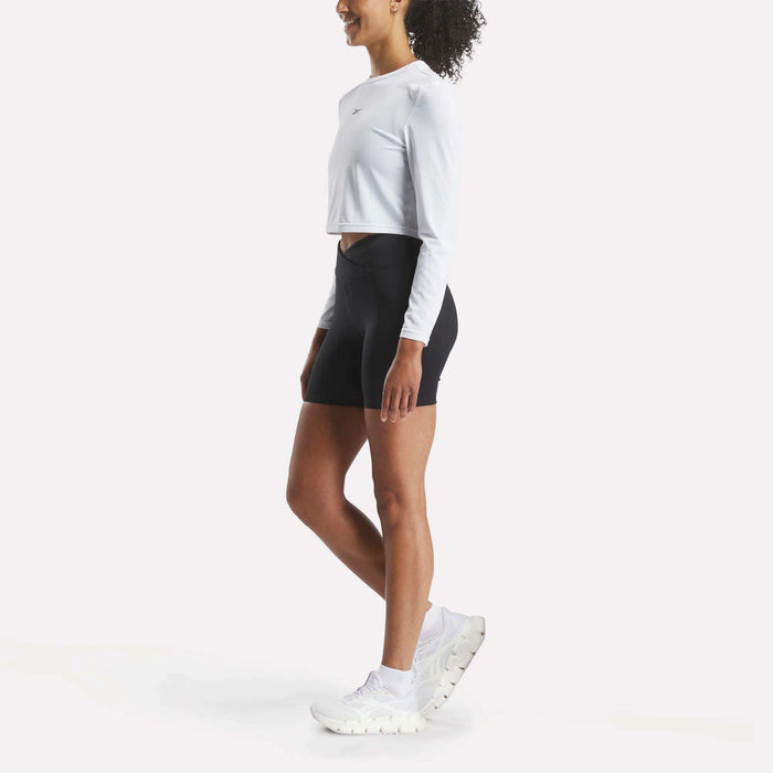 A woman stands sideways against a plain white background, wearing a long-sleeve white crop top, Reebok Women's Core High-Rise Cycle Shorts, and white athletic shoes.