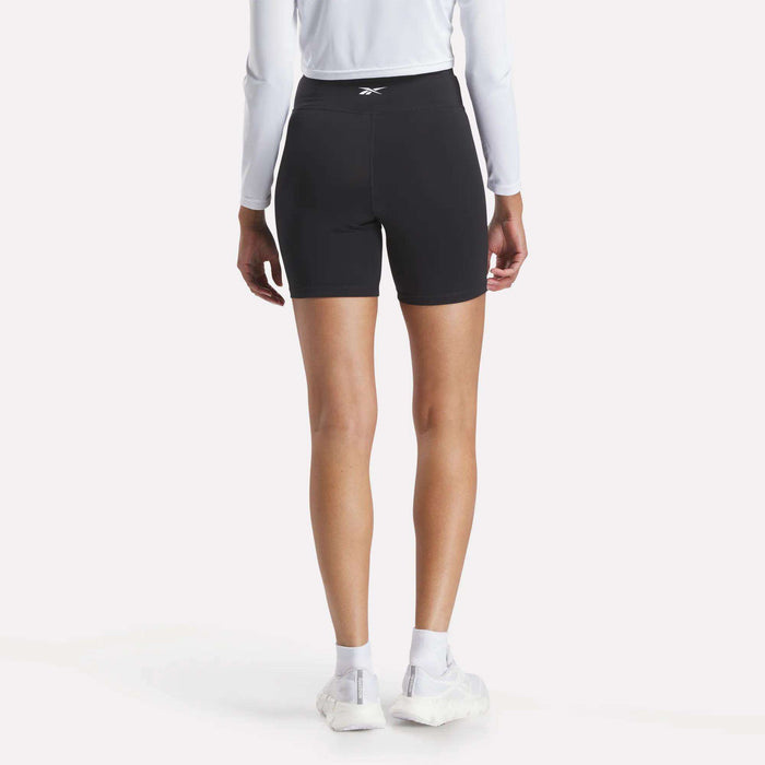 A person in Reebok Women's Core High-Rise Cycle Shorts, a long-sleeve white shirt, white socks, and white sneakers stands with their back to the camera against a plain light background.