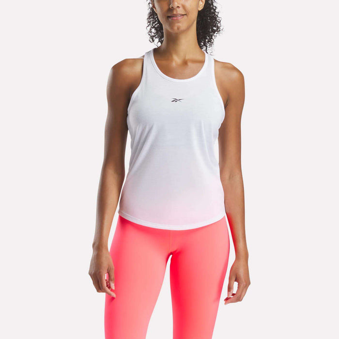 A woman stands facing forward against a plain background, wearing a white Reebok Women's Core Tank Top with moisture-wicking fabric and a small chest logo, paired with bright pink leggings.