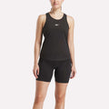 A person wears a black Reebok Women's Core Tank Top and fitted shorts, standing against a plain light background. The tank top has sweat-wicking fabric and a small white Reebok logo on the chest.