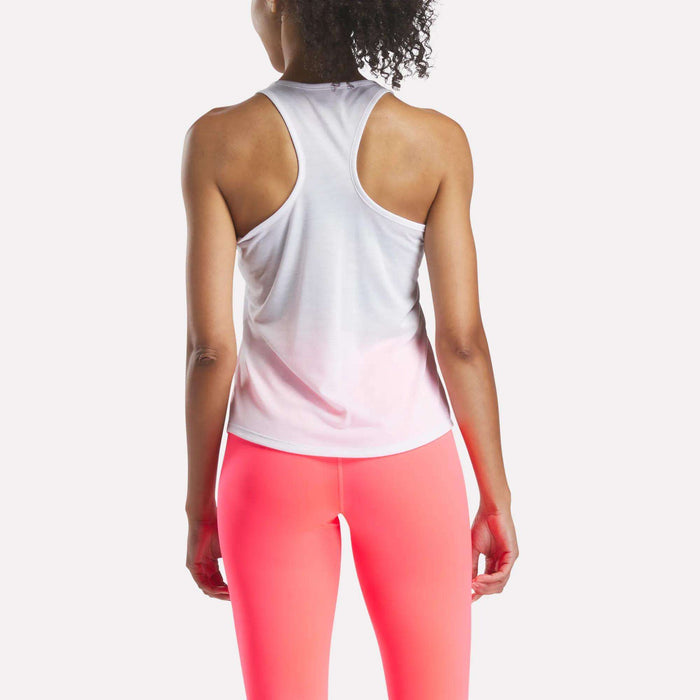 A woman is seen from behind wearing a white Reebok Women's Core Tank Top and bright pink leggings, standing against a plain light background.