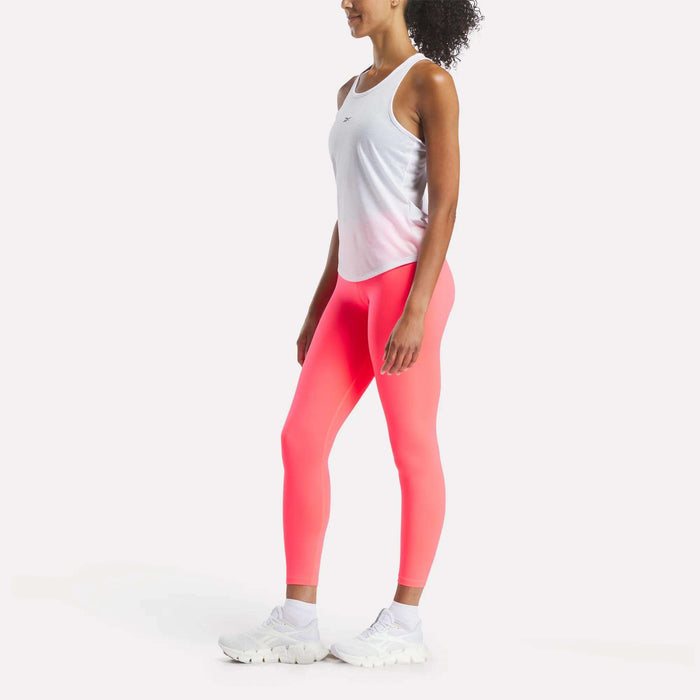 A woman with her hair tied back stands sideways against a plain light background, wearing a Reebok Women's Core Tank Top, bright pink leggings, and white sneakers. Her arms are relaxed at her sides.