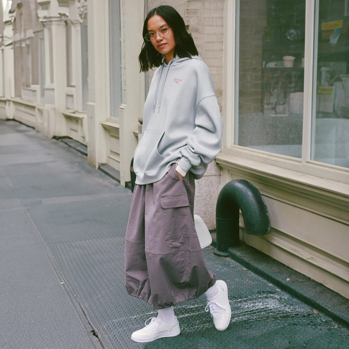 A woman stands on a city sidewalk wearing a light gray oversized hoodie, loose mauve cargo pants, white socks, and white sneakers, with storefront windows and a building facade in the background.