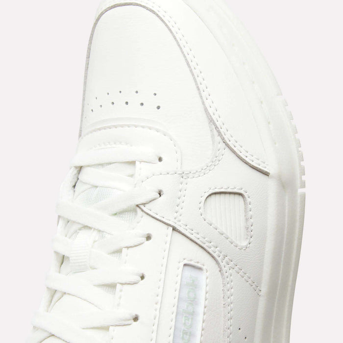 Close-up of a white women’s court shoe—specifically the Reebok Women’s Court LXS Shoes—crafted from full grain leather with detailed stitching, perforations, and white laces, showcasing a modern, minimalist design.