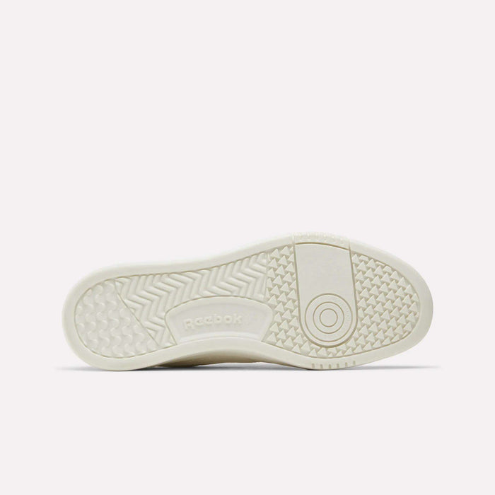A white Reebok sneaker sole is shown, featuring a mix of patterns: zigzags, small raised diamonds, and a circular design near the ball of the foot. The Reebok logo is embossed in the center. Crafted for performance and style, these women’s court shoes—Reebok Court LXS Shoes—offer durability and support whether you’re on or off the court.