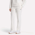 A woman wears Reebok Women's Cozy Classic Sweatpants in off-white, paired with a matching sweatshirt and white sneakers, standing against a light background.
