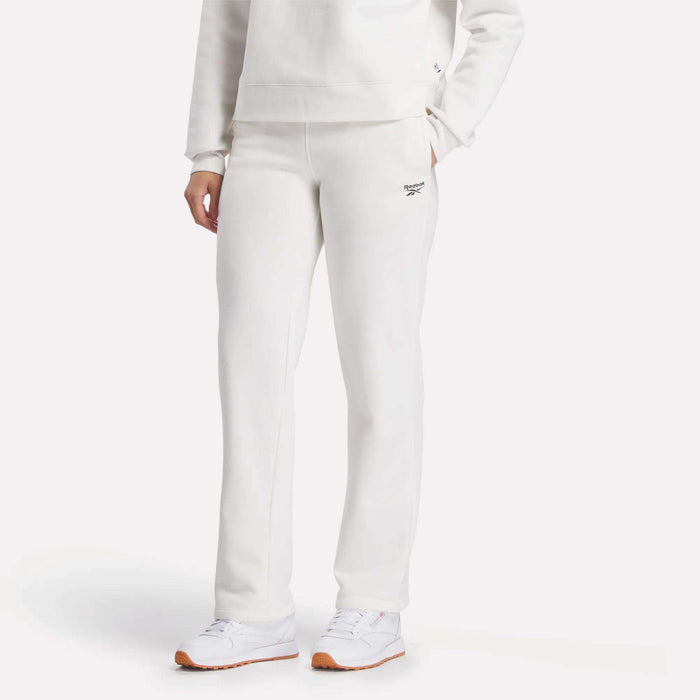 A woman wears Reebok Women's Cozy Classic Sweatpants in off-white, paired with a matching sweatshirt and white sneakers, standing against a light background.
