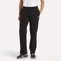 A person wearing Reebok Women's Cozy Classic Sweatpants in black, paired with a white t-shirt and black Reebok sneakers, stands with one hand in their pocket against a plain white background.