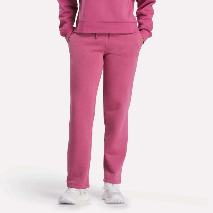 Visible from the shoulders down, a person models Reebok Women's Cozy Classic Sweatpants in bright pink with a matching sweatshirt and white sneakers, standing hands-in-pockets against a plain white background.