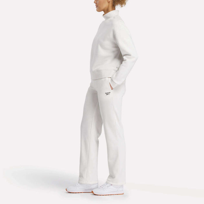A person stands in profile against a white background, wearing Reebok Women's Cozy Classic Sweatpants in white with a matching high-neck sweatshirt and white athletic shoes.