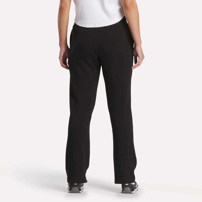 A woman is shown from the back in Reebok Women's Cozy Classic Sweatpants, a white short-sleeve top, and black athletic shoes, standing against a plain light background.