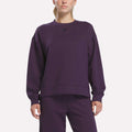 A woman stands against a plain white background wearing matching Reebok apparel in Phantom Purple. She is dressed in the Women’s Crewneck Sweatshirt by Reebok, which features long sleeves, a relaxed fit, and is crafted from comfortable doubleknit fabric.