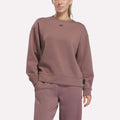 A person wearing a Reebok Women’s Crewneck Sweatshirt in Light Cocoa, along with matching pants, stands with one hand in their pocket against a plain light background.