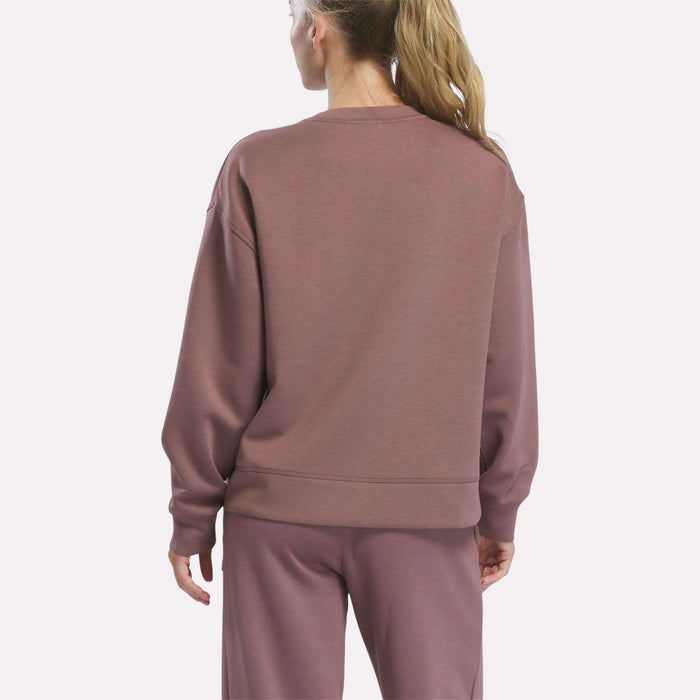 A person with long blonde hair in a ponytail is seen from behind, wearing the Reebok Women’s Crewneck Sweatshirt and matching pants in mauve, standing against a plain background.