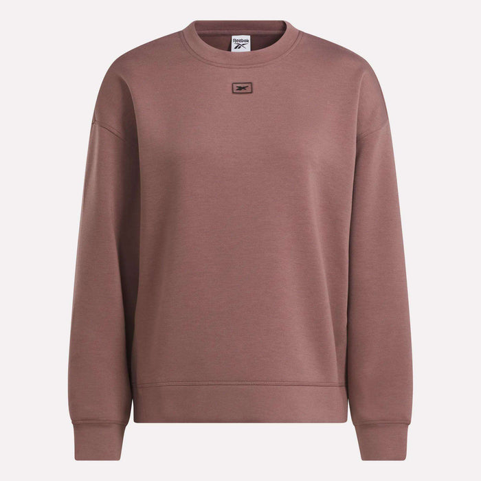 The Women’s Crewneck Sweatshirt by Reebok showcases a classic brown design for women, featuring long sleeves, ribbed cuffs, and a small rectangular logo patch on the upper chest. Made from soft doubleknit fabric, it delivers both comfort and style when worn against a plain, light-colored background.