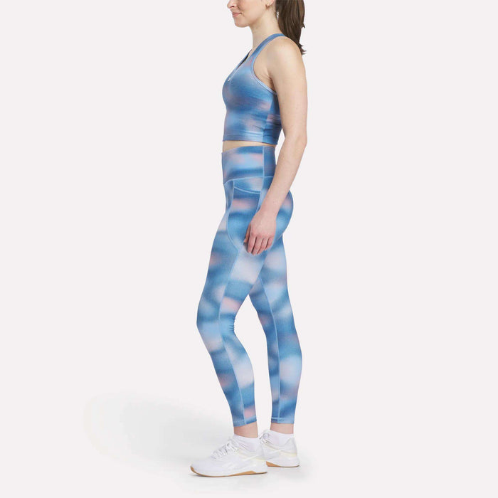 A woman in profile wears a Reebok Women's Cropped Bra Tank Top and matching blue-pink patterned leggings with light-impact support, paired with white athletic shoes, all set against a plain white background.