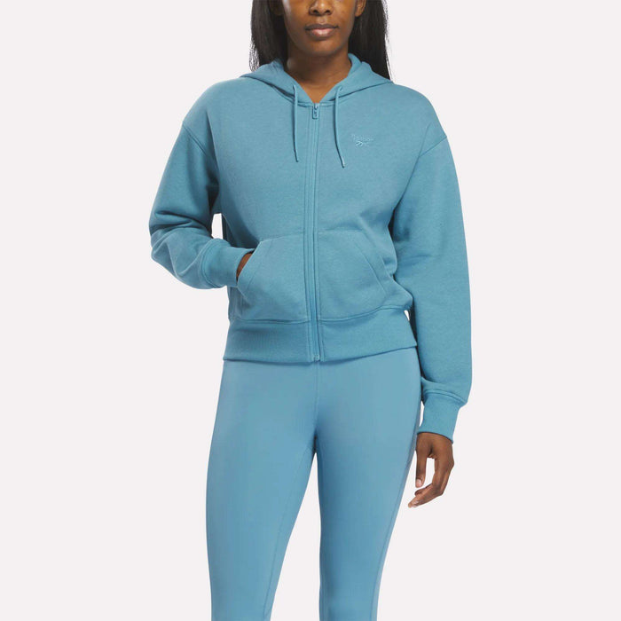 A person wears a Reebok Women's Cropped Full-Zip Hoodie in Warped Blue with matching leggings, both featuring front pockets, standing against a plain white background.