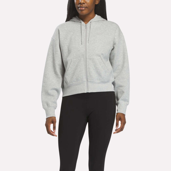 A person wears a Reebok Women's Cropped Full-Zip Hoodie in Grey Heather, featuring front pockets, paired with black leggings against a plain light background.