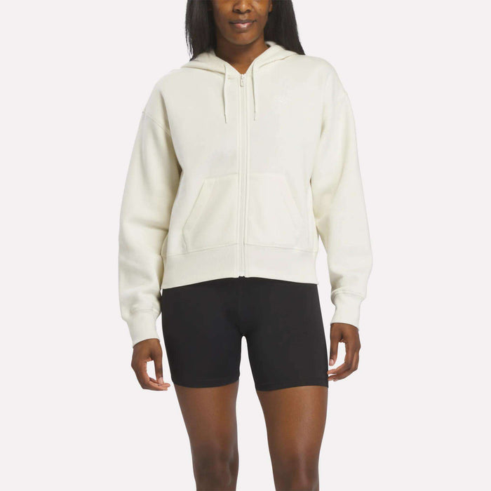 A person wears a Reebok Women's Cropped Full-Zip Hoodie in Alabaster with black fitted shorts, standing against a plain white background.