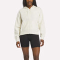 A person wears a Reebok Women's Cropped Full-Zip Hoodie in Alabaster with black fitted shorts, standing against a plain white background.