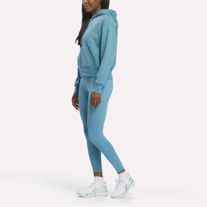 A woman faces left in a Reebok Women's Cropped Full-Zip Hoodie, styled with matching blue leggings and white sneakers, set against a plain white background.