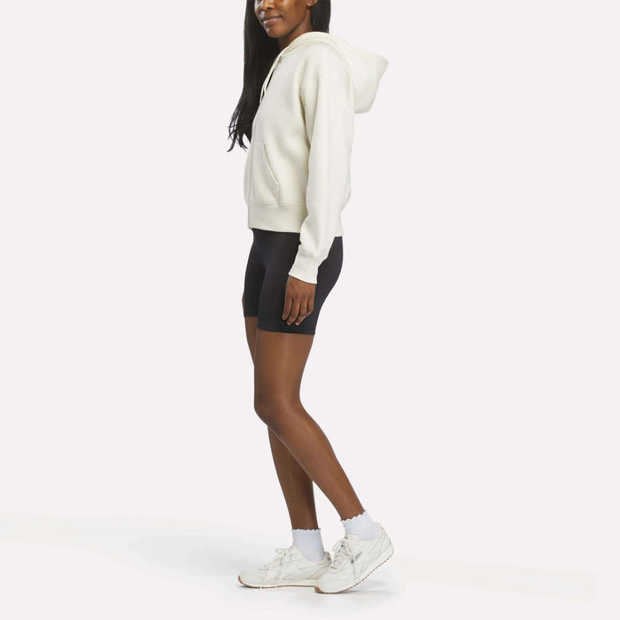 A woman stands sideways against a white background, wearing a Reebok Women's Cropped Full-Zip Hoodie, black biker shorts, white socks, and white sneakers.