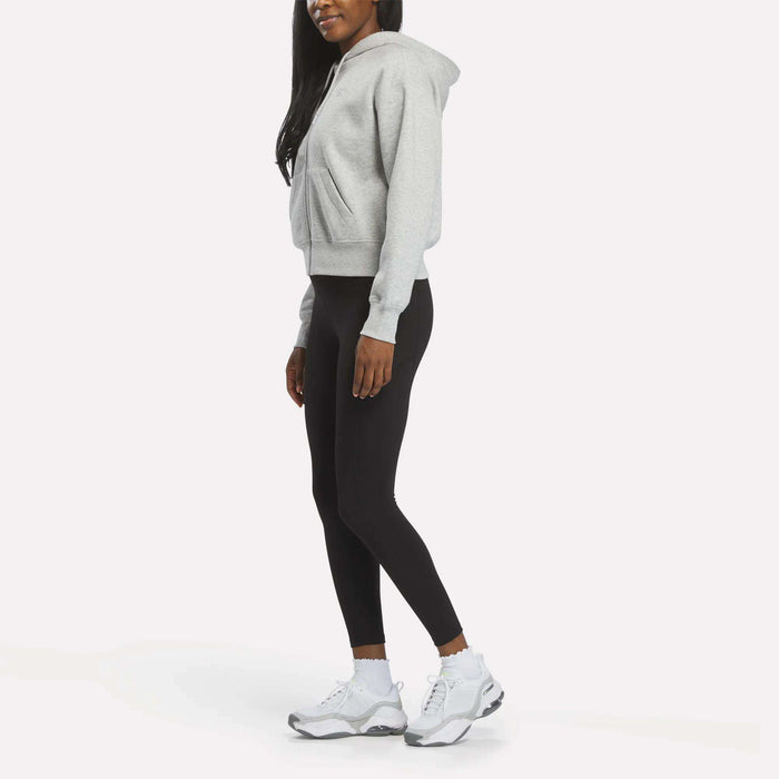 A woman models the Reebok Women's Cropped Full-Zip Hoodie in gray, paired with black leggings and white sneakers, standing sideways against a white background with her hands relaxed at her sides and her face partially out of frame.