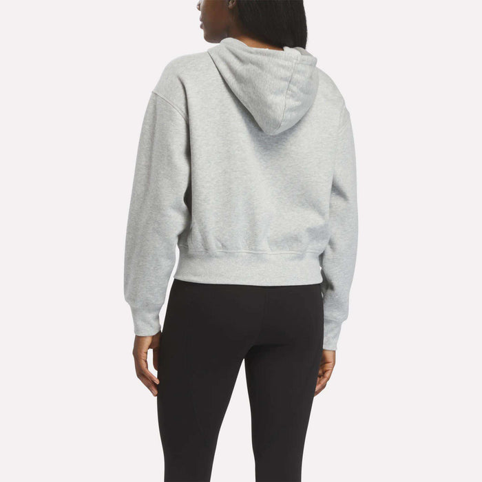 A woman is seen from the back, wearing a light gray Reebok Women's Cropped Full-Zip Hoodie and black leggings, standing against a plain white background.