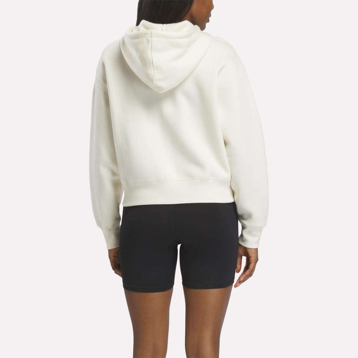A person facing away wears a white Reebok Women's Cropped Full-Zip Hoodie with an embroidered logo and black fitted shorts, standing against a plain light background.