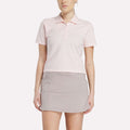 A woman stands facing forward against a white background, wearing a Reebok Women's Fitted Polo Shirt in light pink and a light gray tennis skirt.