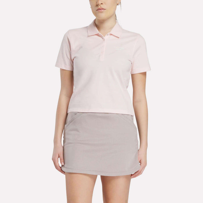 A woman stands facing forward against a white background, wearing a Reebok Women's Fitted Polo Shirt in light pink and a light gray tennis skirt.