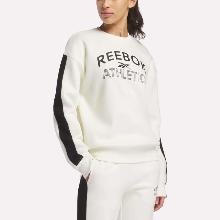 A woman wears a Reebok Women's Fleece Crew Sweatshirt in Chalk, featuring REEBOK ATHLETIC in black, paired with white pants with black side stripes. The soft brushback fleece offers relaxed, stylish comfort.