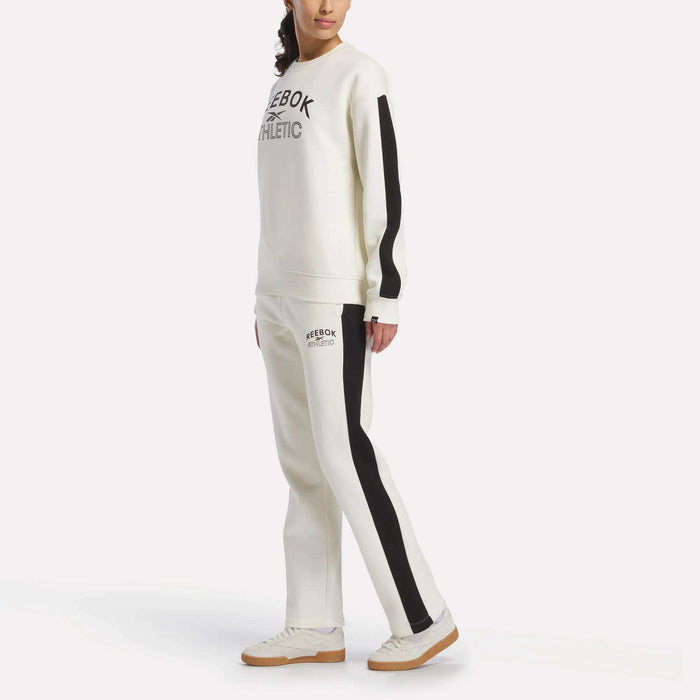 A woman stands sideways in a Reebok Women's Fleece Crew Sweatshirt and matching sweatpants with black side stripes and beige sneakers. The cozy brushback fleece enhances the look, with only part of her face visible.