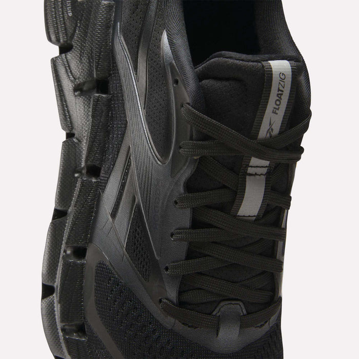 Close-up of a black Reebok Women's FloatZig 2 Running Shoe with energy-returning cushioning, mesh upper, black laces, zigzag-patterned sole, and a white and black tongue label reading ZIG KINETICA II FLOATZIG.