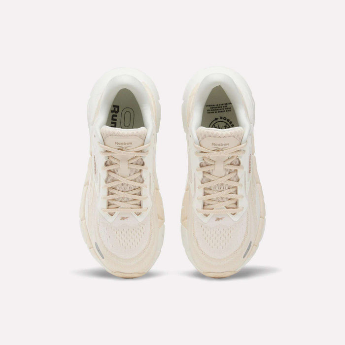 A pair of Women's FloatZig Double Running Shoes by Reebok in beige are shown from above, featuring laces, mesh texture, and logo details on the tongues—designed for max-cushioned runs—set against a plain white background.