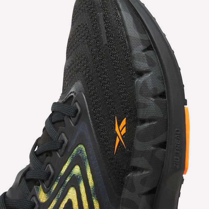 Close-up of Reebok Women's FloatZig Tread Running Shoes with a breathable mesh upper, yellow and green accents, orange logo, and a rugged black/camouflage sole. Features an orange heel detail. Ideal for treadmill workouts.