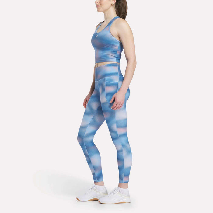 A woman stands in profile wearing Reebok Women's Fluid Print High-Rise Tights with a matching sports bra and white sneakers, highlighting the high-rise waist against a plain white background.